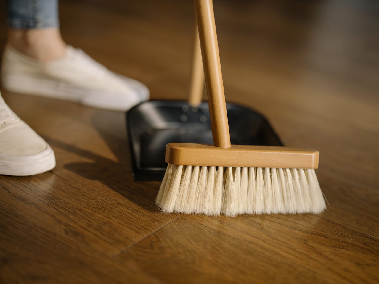 Services-01 Close-up of a broom and dustpan with white sneakers indoors, representing housekeeping and cleanliness.